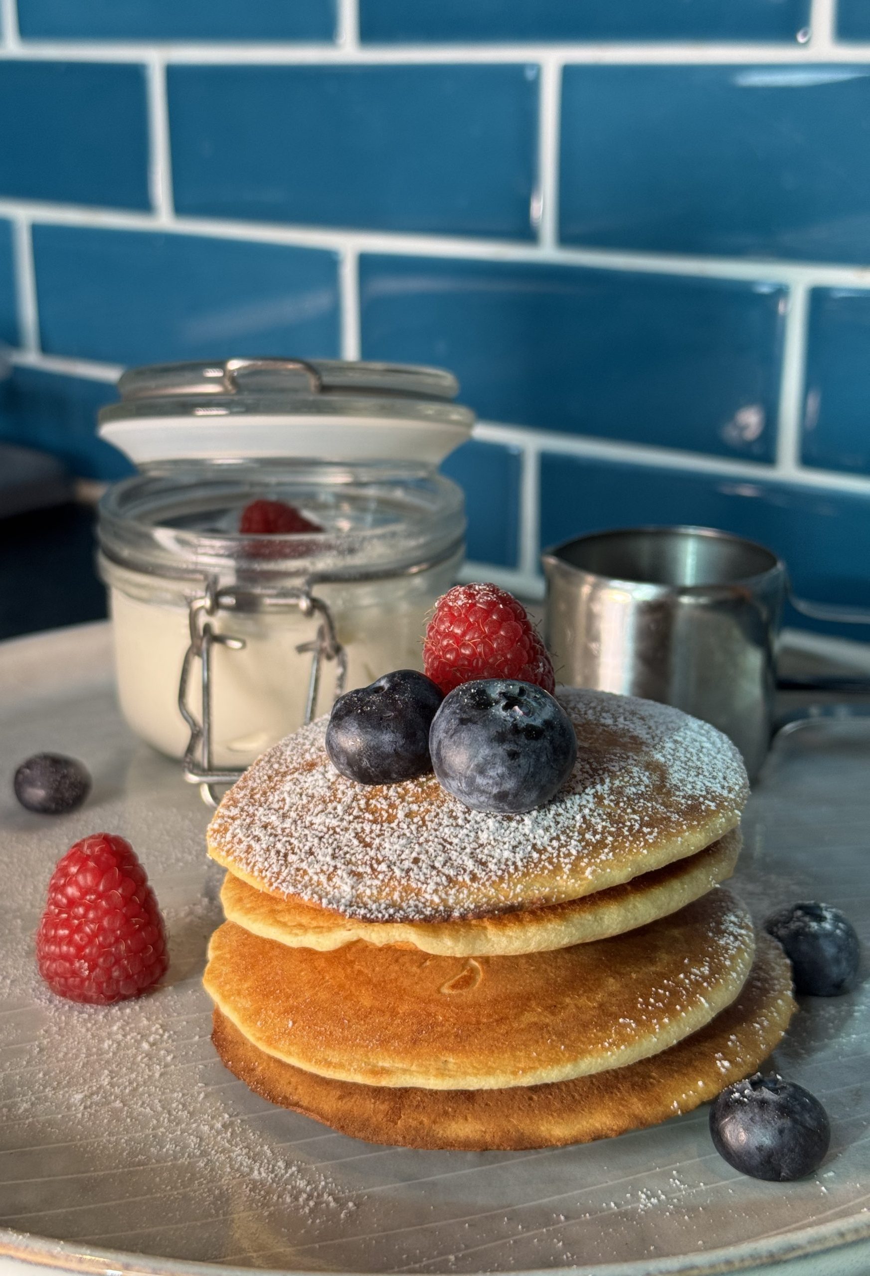 American pancakes mit Puderzucker und Beeren verziert. Dahinter ein Joghurt in einem Glas